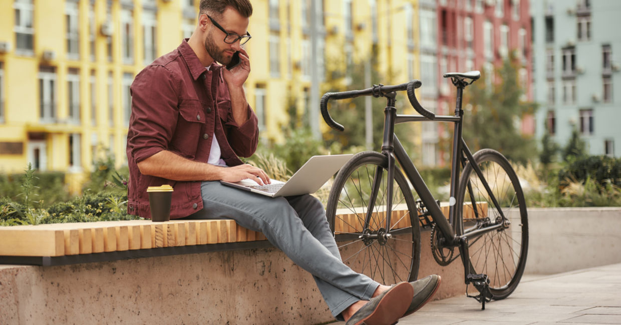 Young man working on laptop and talking by phone while sitting on the bench near his bicycle Young man working on laptop and talking by phone while sitting on the bench near his bicycle