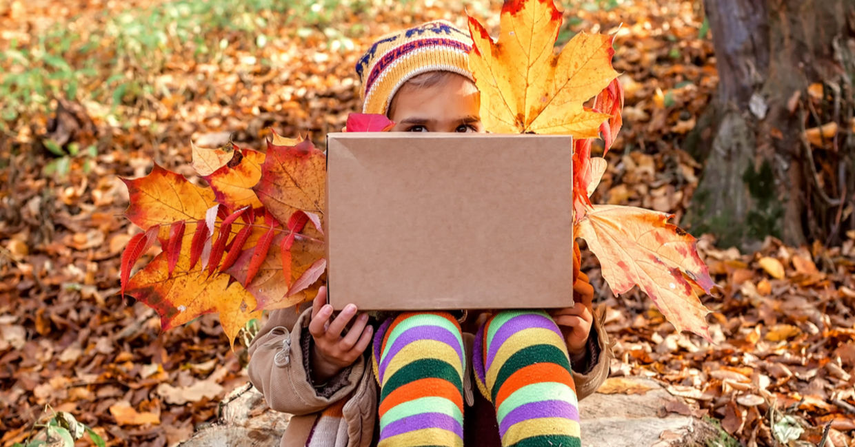 A young girl holding autumn leaves peeks from behind a large gift box. A young girl holding autumn leaves peeks from behind a large gift box.